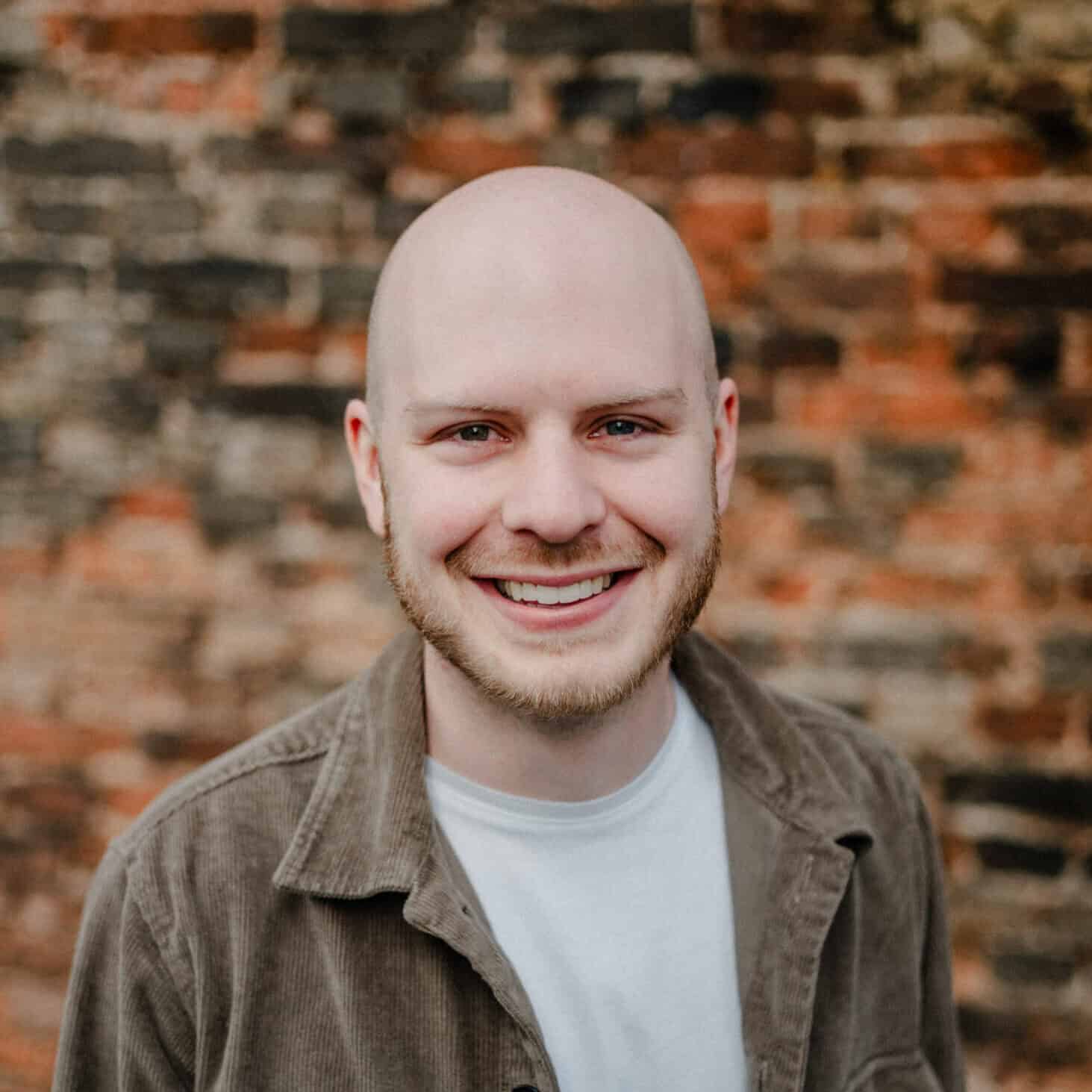 Alex Hintergrund: Ziegelmauer, Porträt eines lächelnden jungen Mannes mit glattem Kopf und Bart, trägt braunen Cordjacke und weißes T-Shirt, professionelles Foto, urbaner Stil, freundliche Ausstrahlung, Lifestyle, Gesichtsausdruck, kreative Aufnahme.