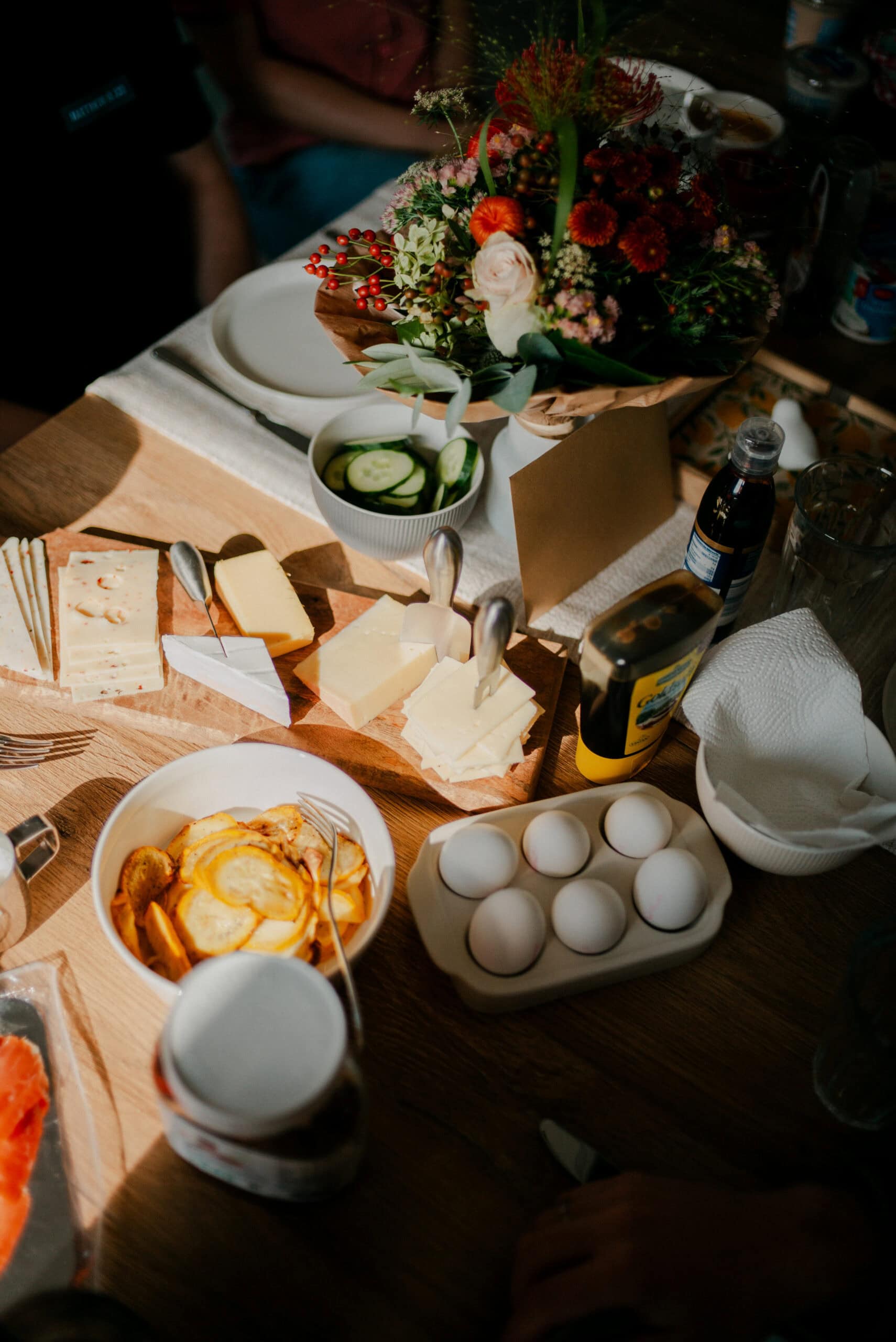 Blumenstrauß auf einem gedeckten Tisch mit Käse, Eiern, Gurkenscheiben und Frühstückszutaten, geselliger Brunch, natürliche Beleuchtung, Mahlzeit, Frühstück, Tischdekoration, romantischer Anlass, Premium-Qualität.