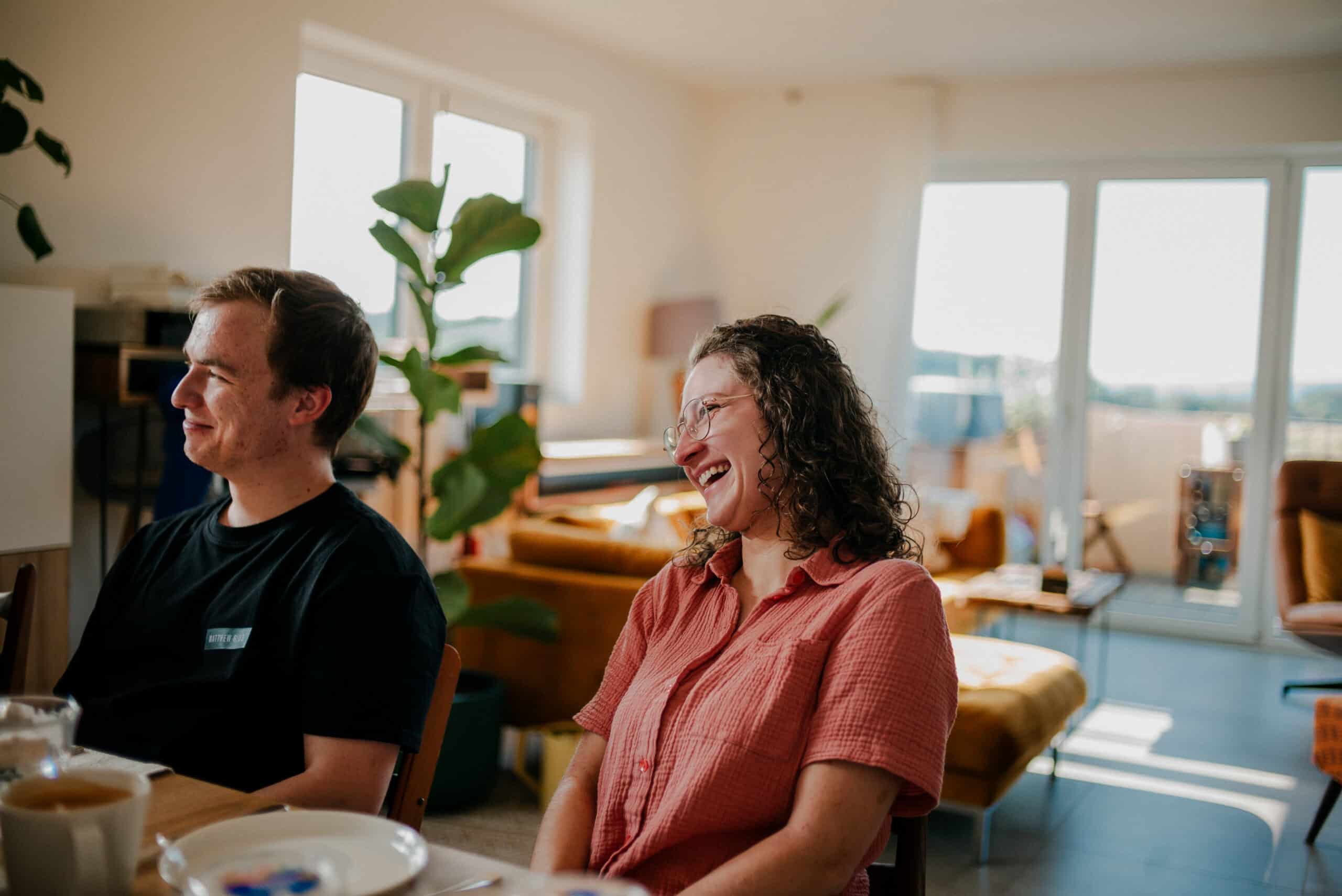 Fröhliche Menschen beim Abendessen in einer hellen, modernen Wohnung mit großen Fenstern, Pflanzen und gemütlicher Einrichtung, Lifestyle und gemeinsames Beisammensein, warmes Licht, entspannte Atmosphäre, Wohnungskonzept.