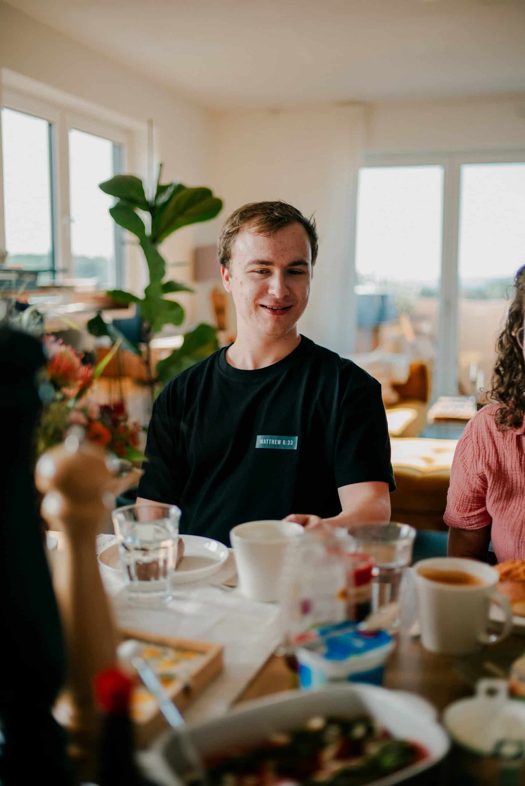 - Fröhlicher junger Mann beim Frühstück in einem hellen, freundlichen Raum, umgeben von Tassen, Tellern und Blumen, symbolisiert Gemeinschaft und Genuss.