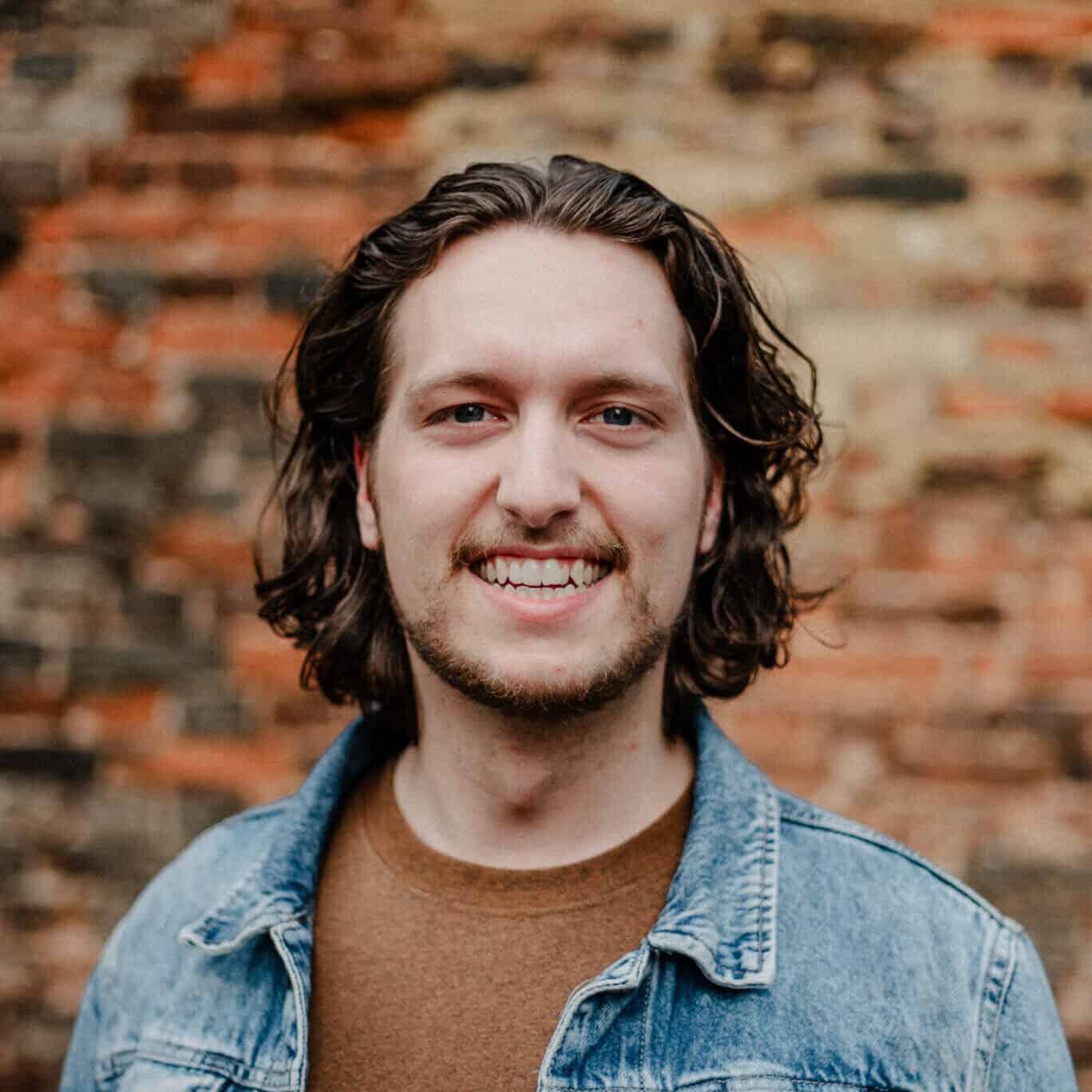 Simon Gesicht eines jungen Mannes mit lockigem braunem Haar vor einer alten Backsteinmauer, lächelnd, im Casual-Look, für Mosaikkirche Bitburg SEO.
