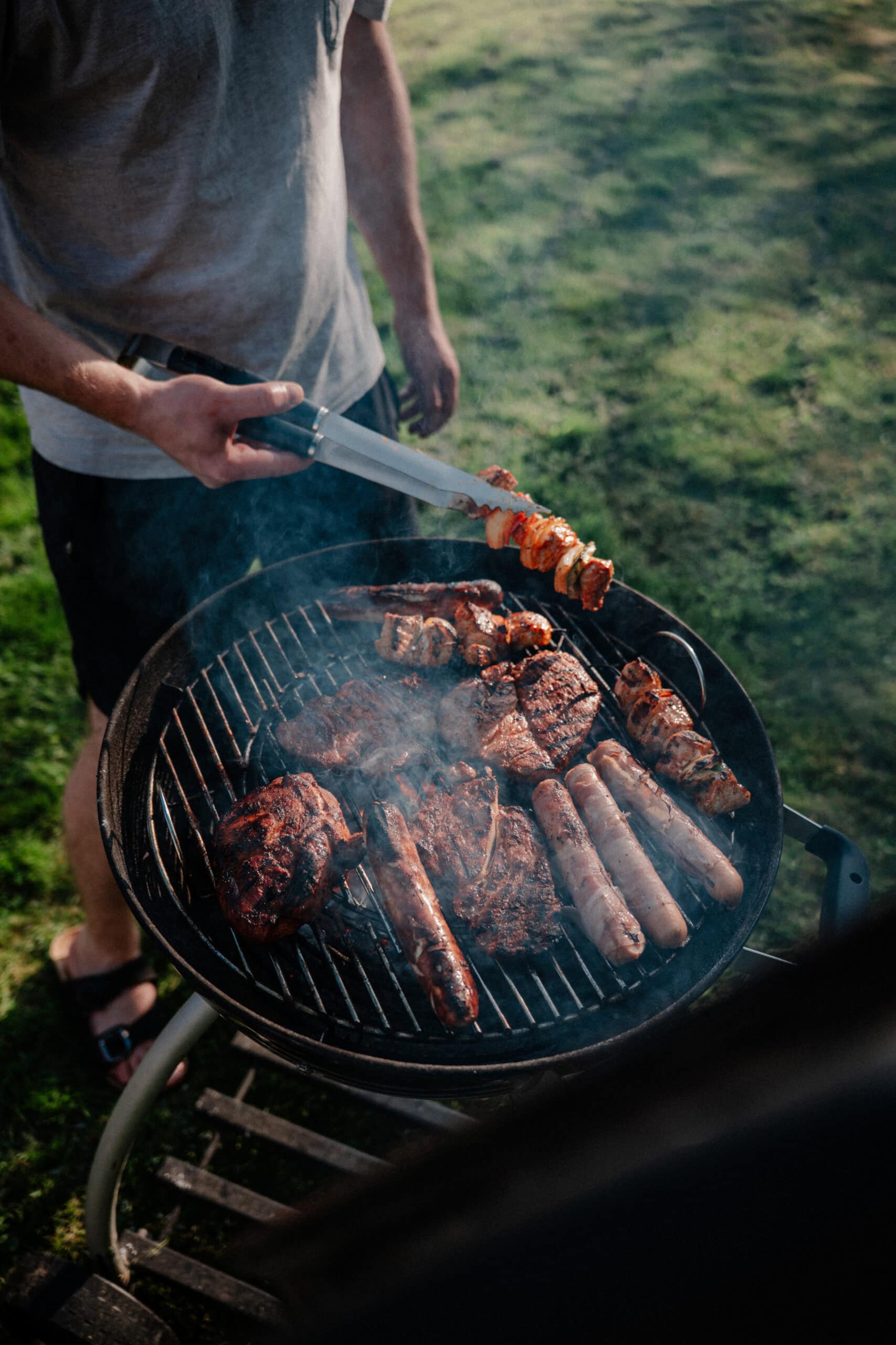 Saftiges Grillen mit verschiedenen Fleischsorten auf einem runden Holzkohlegrill im Freien an einem lauen Sommerabend.