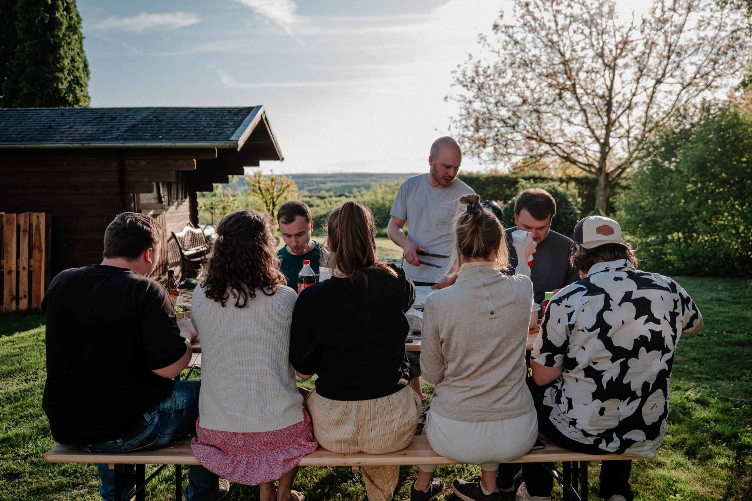 Freiluftgrillabend mit Freunden auf der grünen Wiese vor einer kleinen Holzhütte bei Sonnenuntergang, entspannte Atmosphäre, Gemeinschaft und Natur.